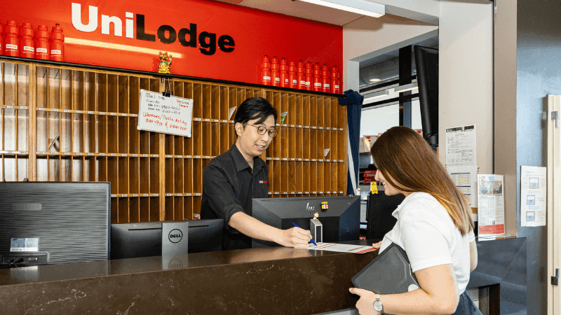 Man assisting woman at StudentLiving Darwin reception desk with red UniLodge sign above.