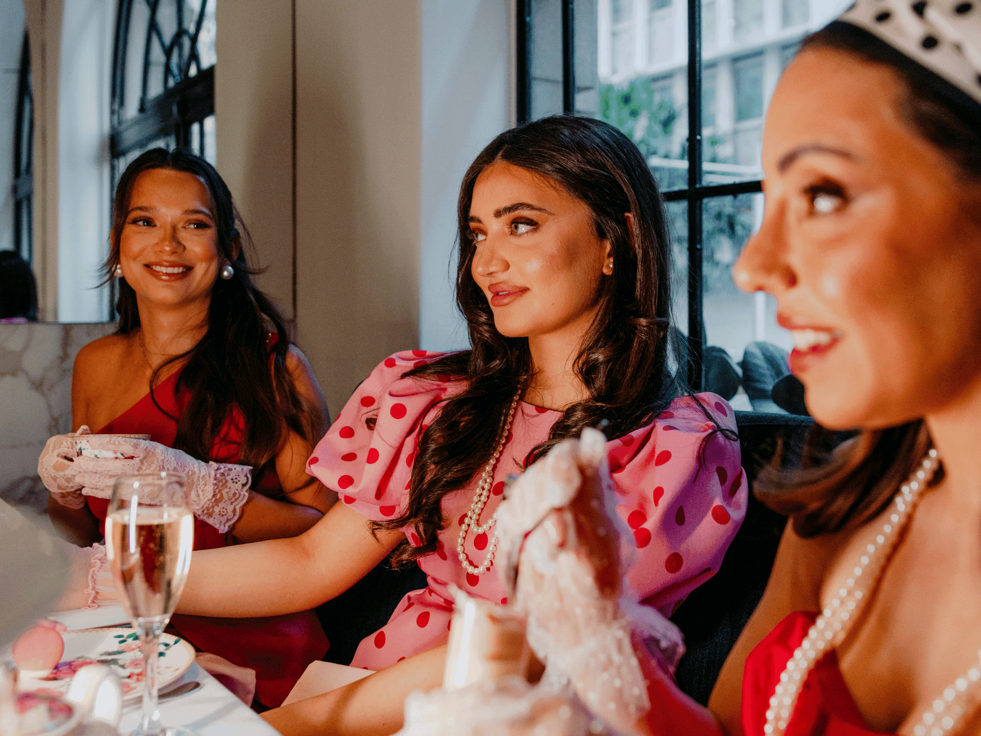 Three women in elegant attire enjoying Group High Tea together with champagne glasses and delicate treats in a bright setting.