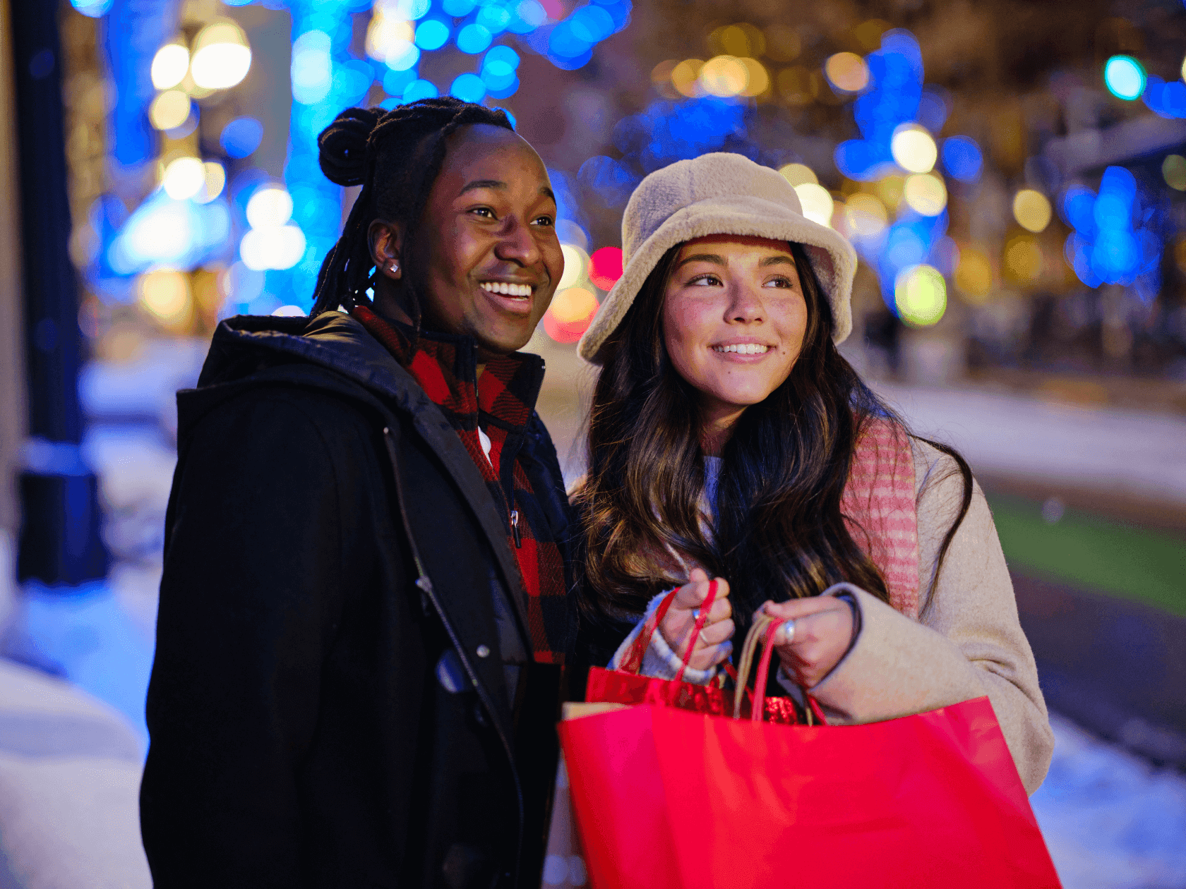 Two women smiling, one with a hat and shopping bags, standing in a snowy area with colorful lights.