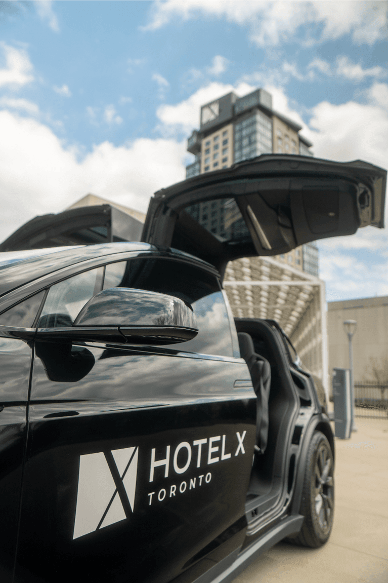 Close-up of a black Tesla at Hotel X Toronto, with an open falcon-wing door by the hotel entrance under a cloudy sky
