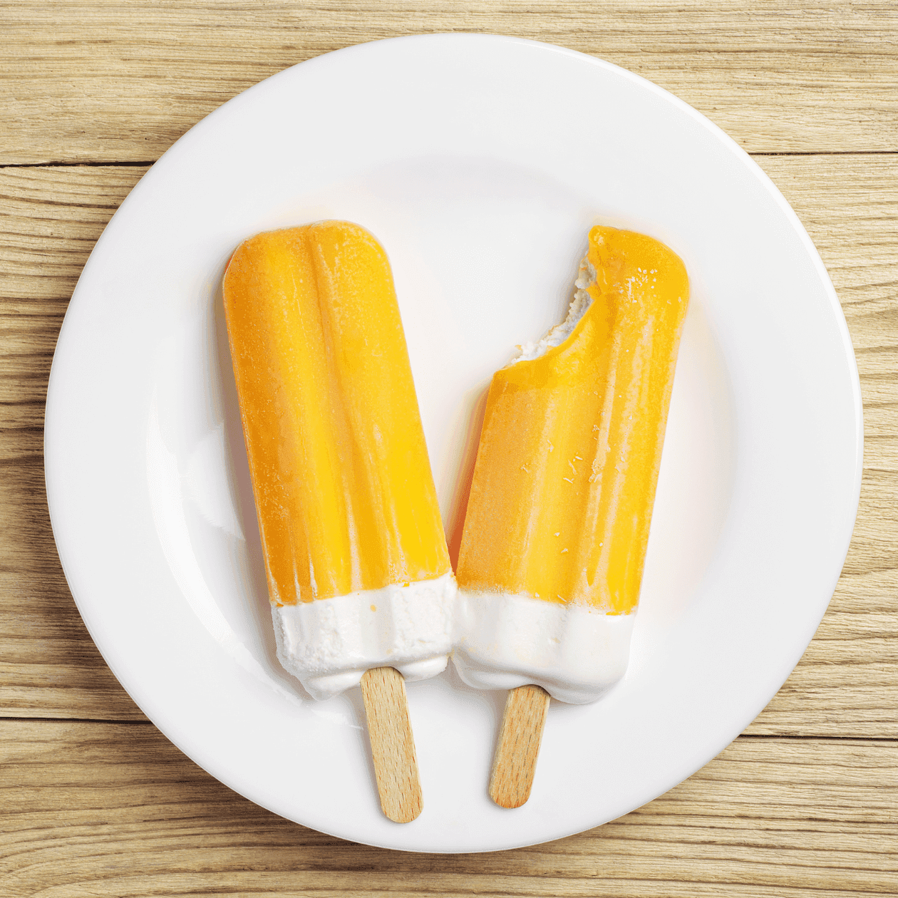 Two popsicles, one bitten, on a white plate atop a wooden surface.