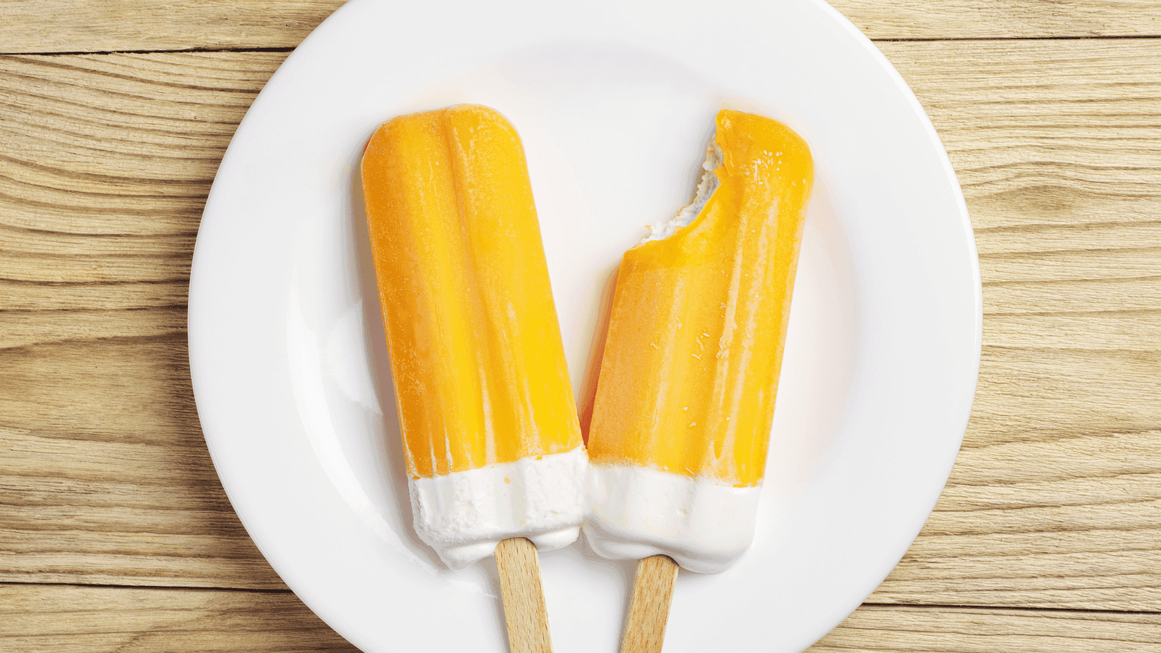 Two popsicles, one bitten, on a white plate atop a wooden surface.