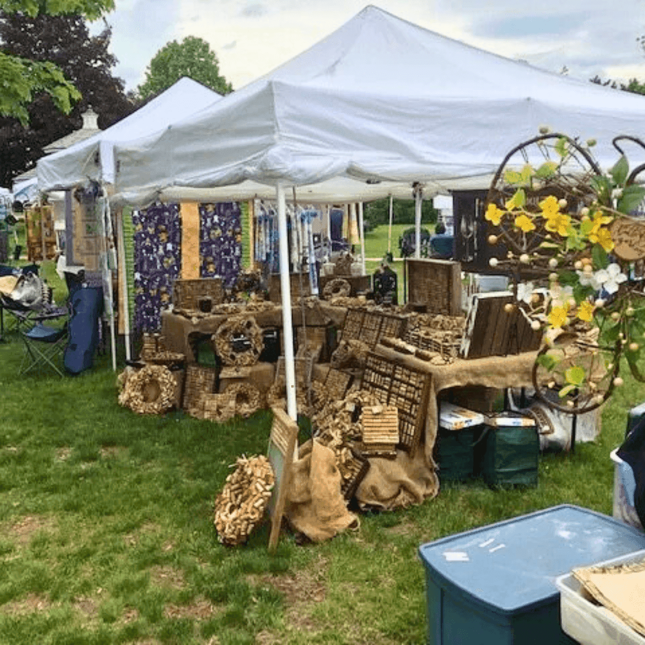 White canopy tent with various goods, storage boxes, and wreaths displayed at an outdoor market on grass.