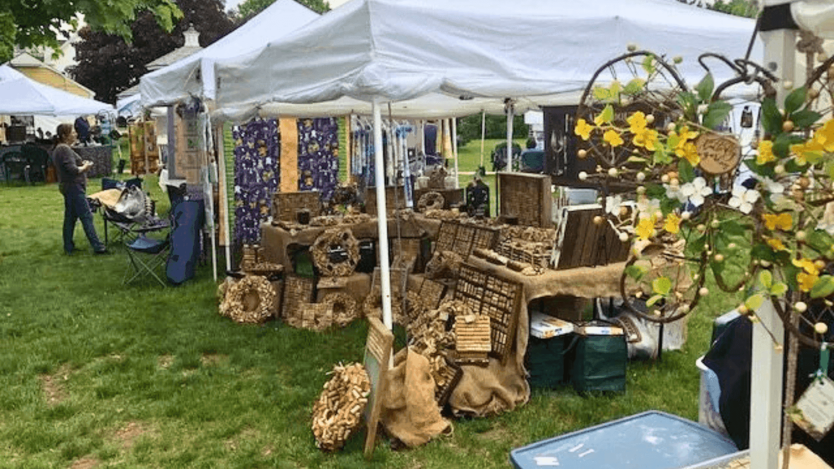 White canopy tent with various goods, storage boxes, and wreaths displayed at an outdoor market on grass.