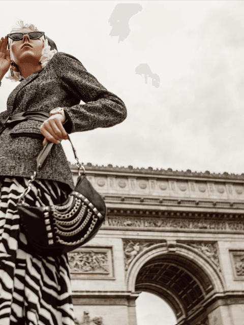Stylish woman in designer outfit and sunglasses poses confidently in front of the Arc de Triomphe in Paris.