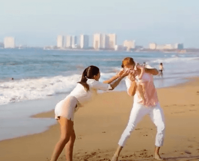 A couple in white clothing holds hands and plays on a sandy beach with ocean waves and distant buildings at Playa Los Arcos Puerto Vallarta 2023