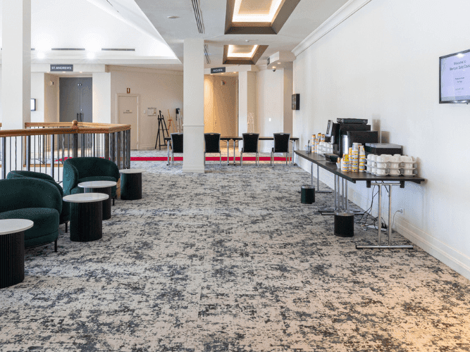 A corridor with green chairs, tables, a monitor on the wall, and a carpeted floor.
