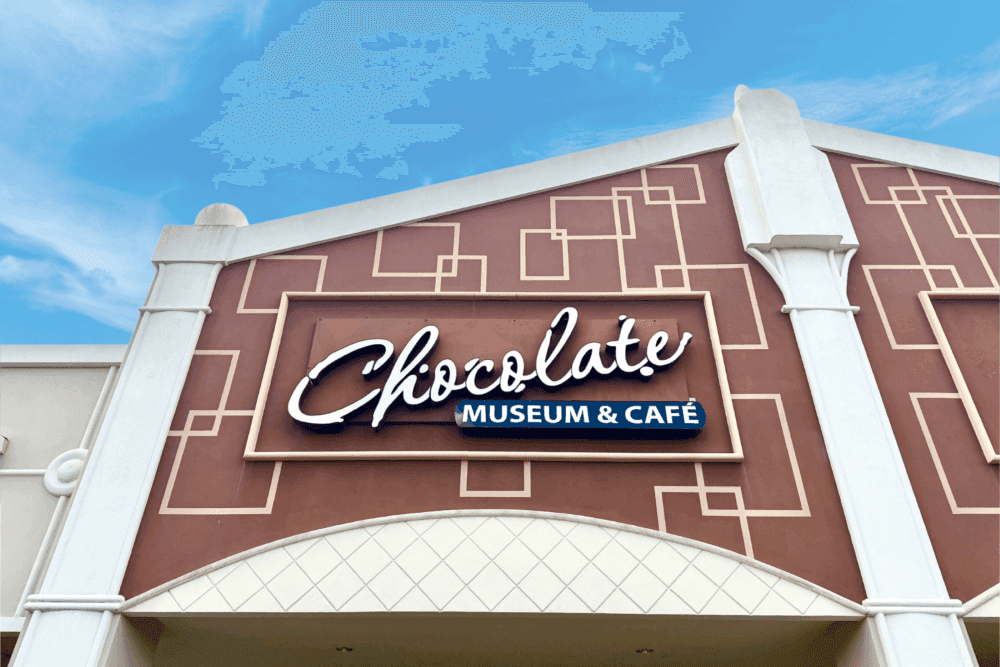 The front of a building with brown plaster and geometric squares and a sign that reads Chocolate Museum and Cafe against a blue sky.