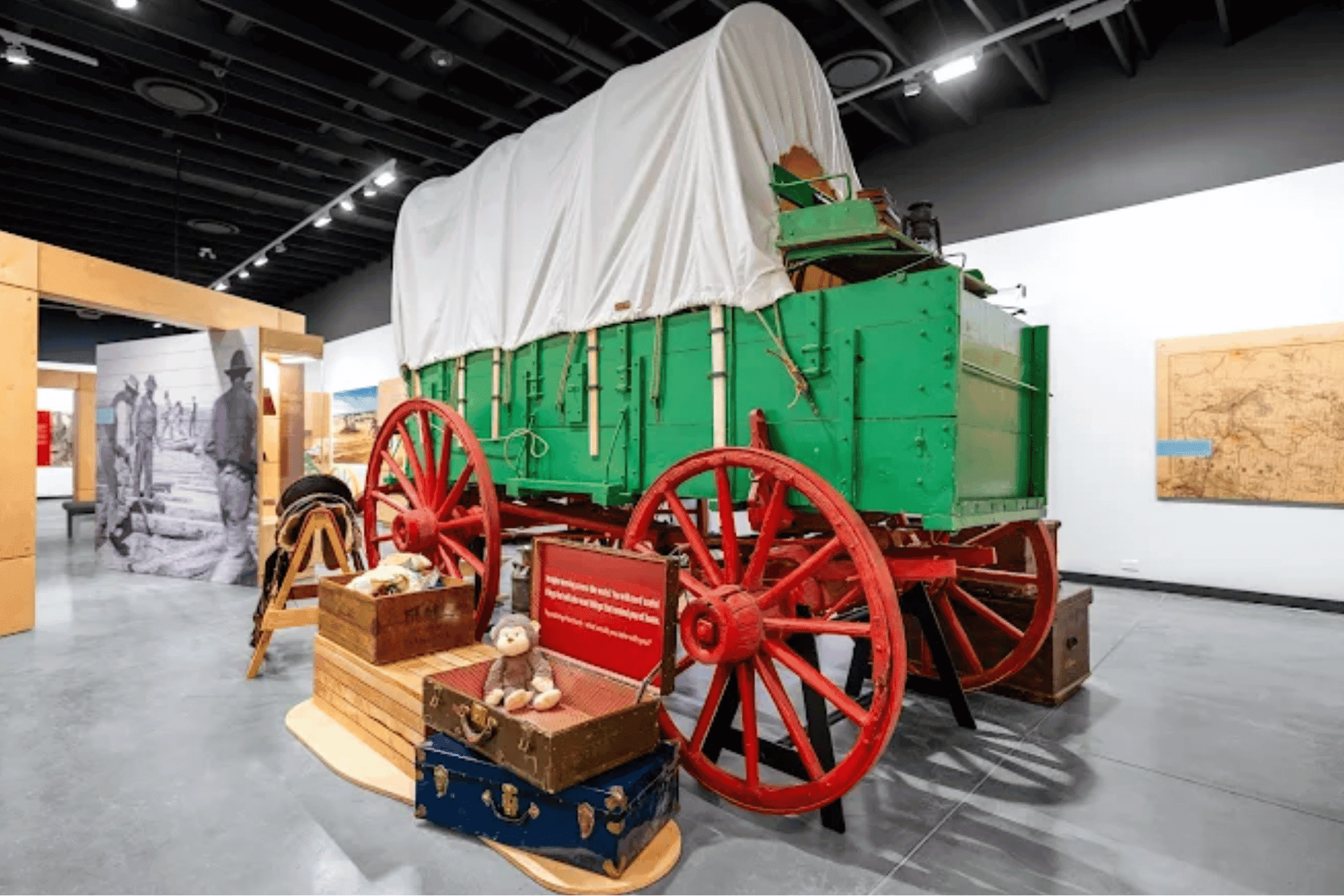 Photograph of a covered wagon exhibit in a museum setting, showcasing a green wagon with large red wheels and a white canvas cover. Surrounding the wagon are vintage suitcases and a teddy bear, highlighting historical travel and pioneer life.
