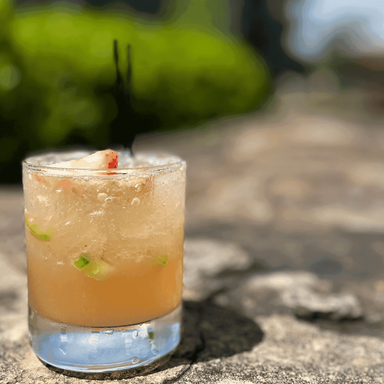 A clear glass with a cocktail, garnished with fruit and herbs, sits on a stone surface.