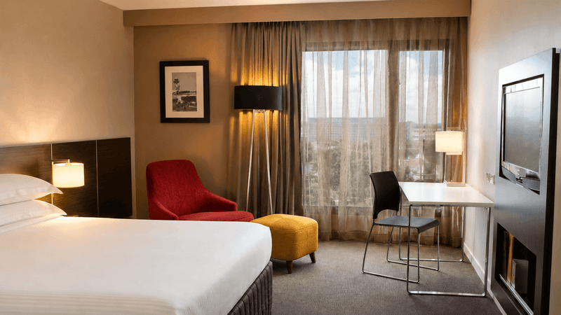 Superior Room with queen bed, work desk, red accent chair, and city views at Novotel Sydney International Airport.