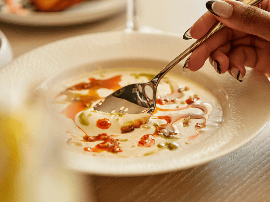 A person’s hand holding a spoon over a bowl of creamy soup with toppings.