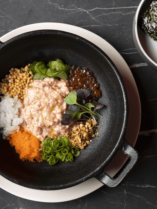Bowl of mixed ingredients including rice, grains, vegetables, and a separate bowl of seaweed on a black surface.