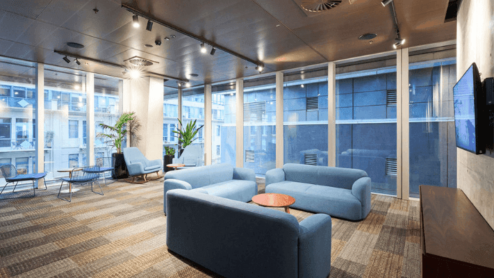 Modern lounge with blue couches, large windows, and a mounted TV.