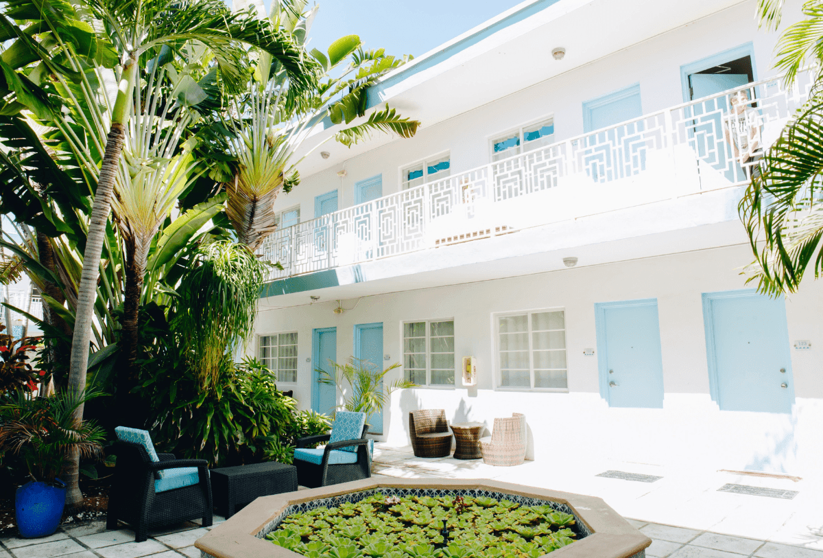 A white building with blue doors and balconies, surrounded by palm trees and a garden with a small pool.