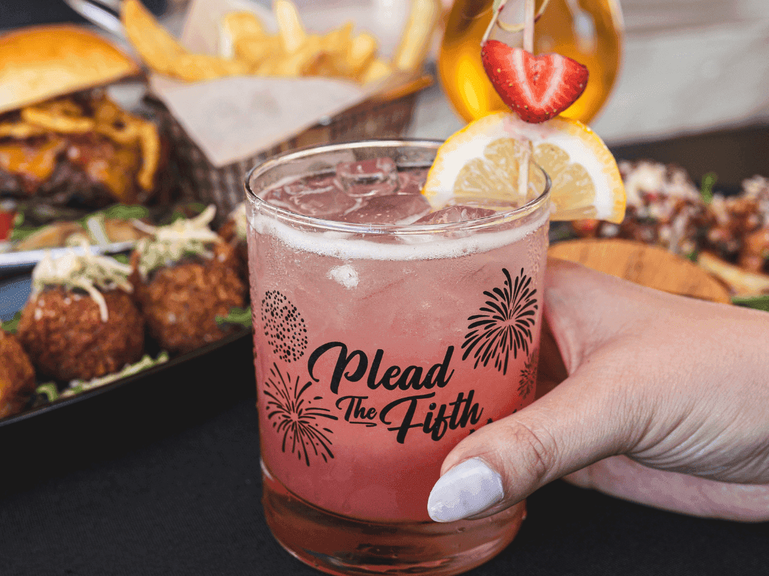 Hand holding signature cocktail with food in background
