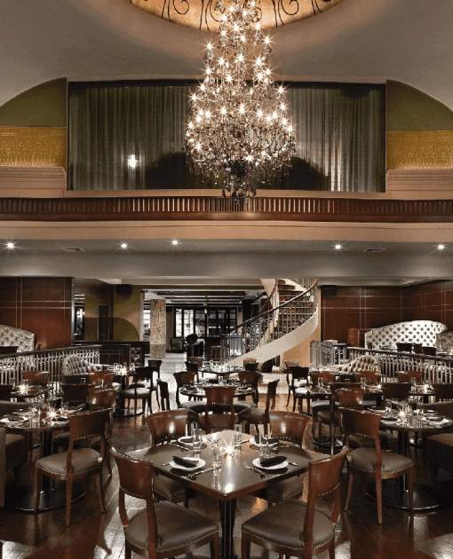 Dining tables facing curved staircase under grand chandelier beside upper balcony seating area at The Kimberly Hotel