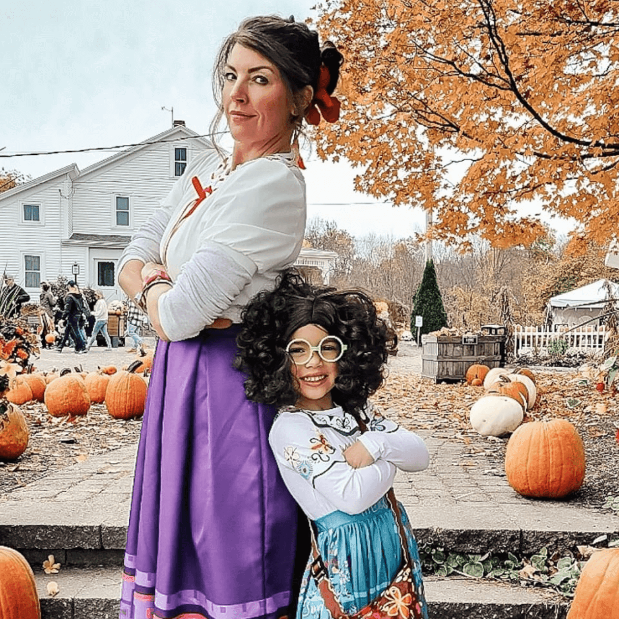 Woman and child in costumes pose on steps by pumpkins and autumn leaves in front of a house.