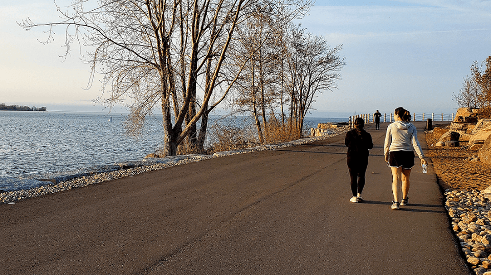 Two women walking along paved waterfront pathway beside bare trees facing the lake water and blue sky at Hotel X Toronto