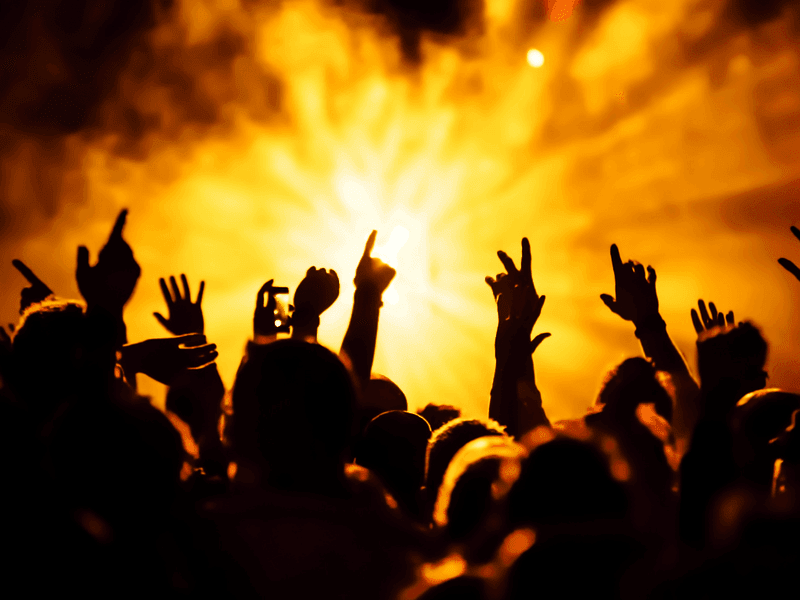 Concert crowd with raised hands silhouetted against intense golden stage lights and atmospheric smoke.