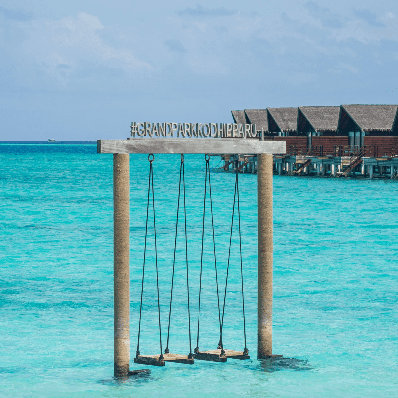 Wooden swing set standing in turquoise lagoon waters with overwater bungalows visible in the background under blue sky at Grand park Kodhipparu, Maldives