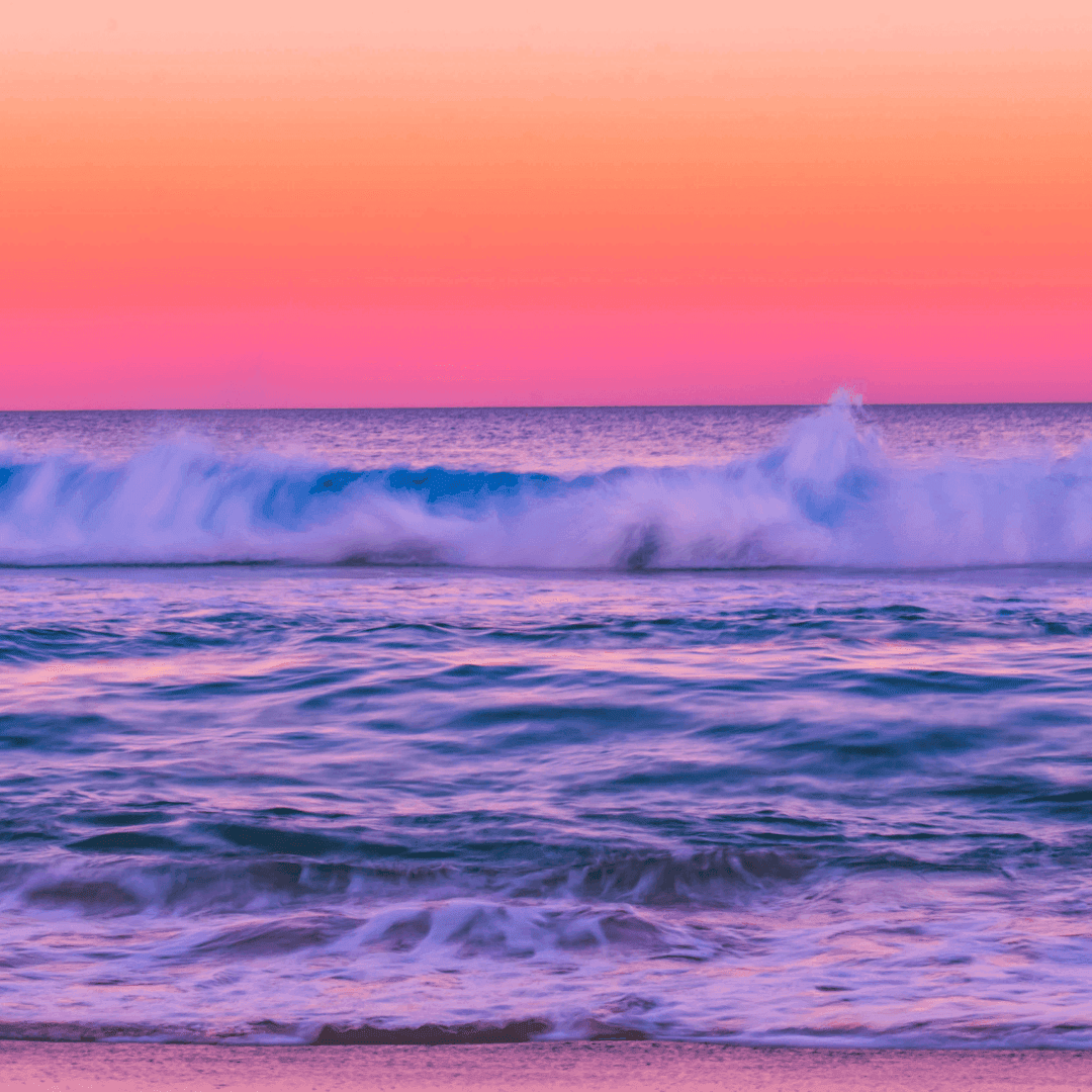 Beautiful beach with gradient orange, pink, blue, and purple sky shining onto the crashing waves hitting the shoreline.