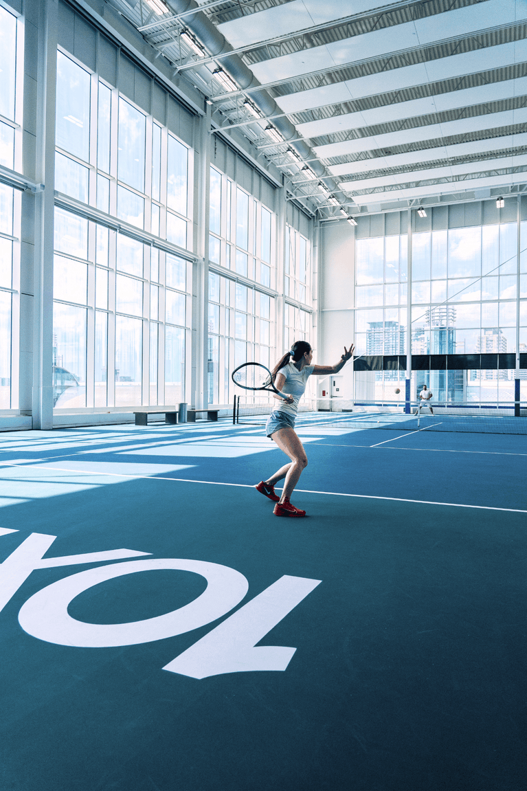 Woman playing tennis on a blue indoor court by floor-to-ceiling windows under a high ceiling at Hotel X Toronto