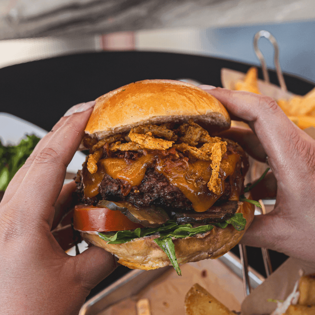 A person holding a burger with cheese, beef, tomato, lettuce, and fried onion.