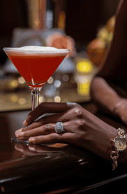 Two cocktails sit on a dark wood bar counter next to a woman\'s hand adorned with a shiny ring & watch at Margaux Restaurant