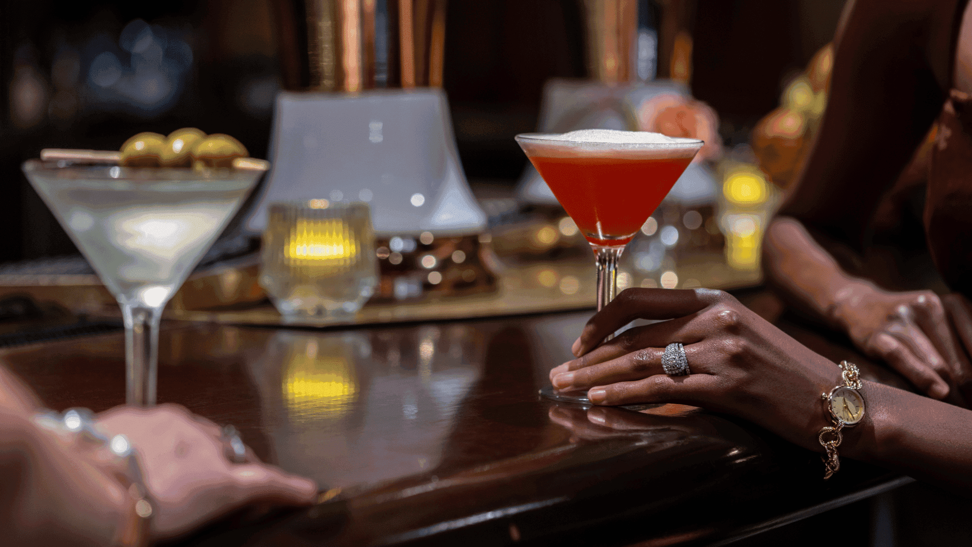 Two cocktails sit on a dark wood bar counter next to a woman\'s hand adorned with a shiny ring & watch at Margaux Restaurant