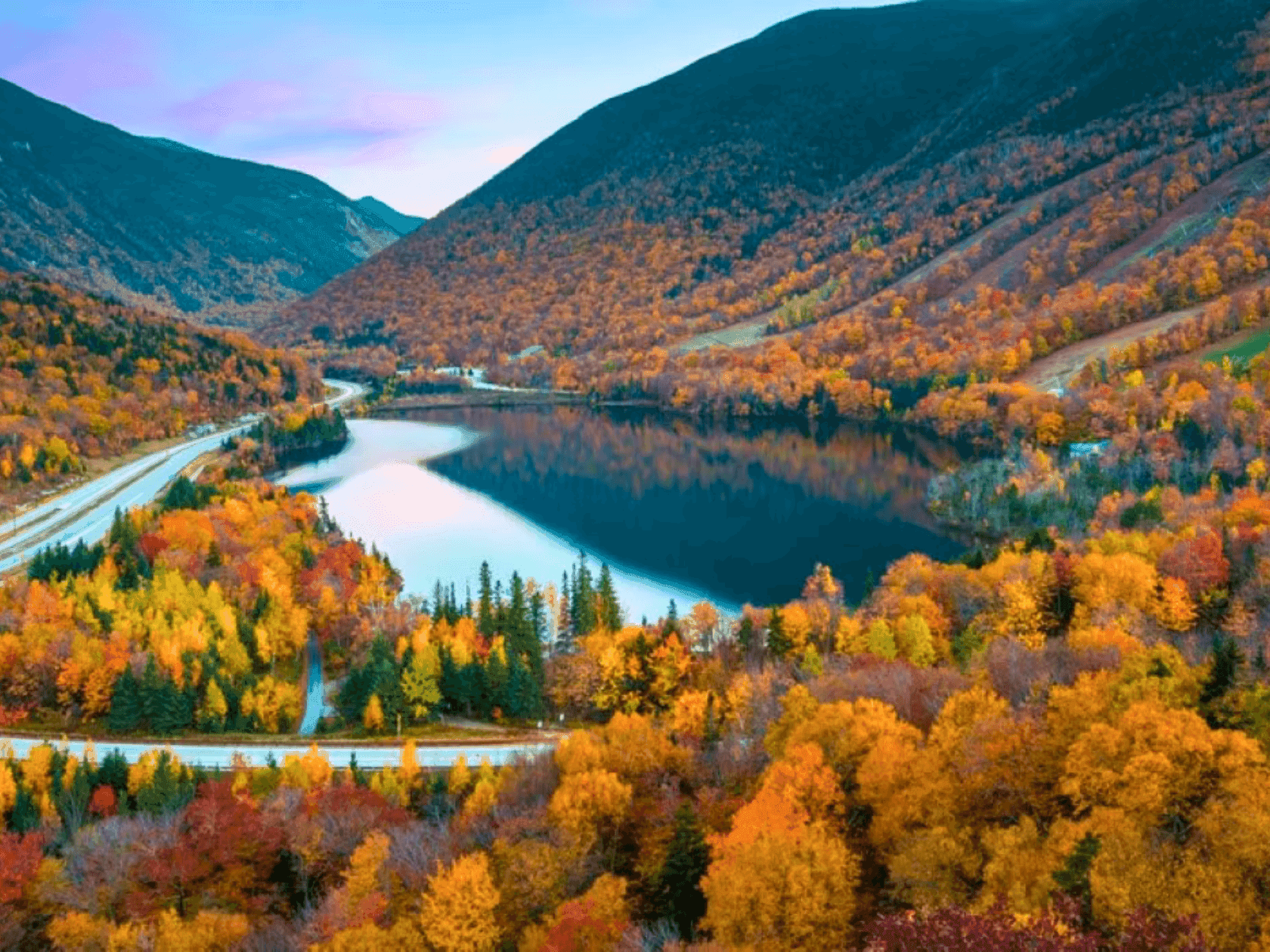 Aerial view of a lake with autumn-colored trees and a winding road in a mountainous valley.