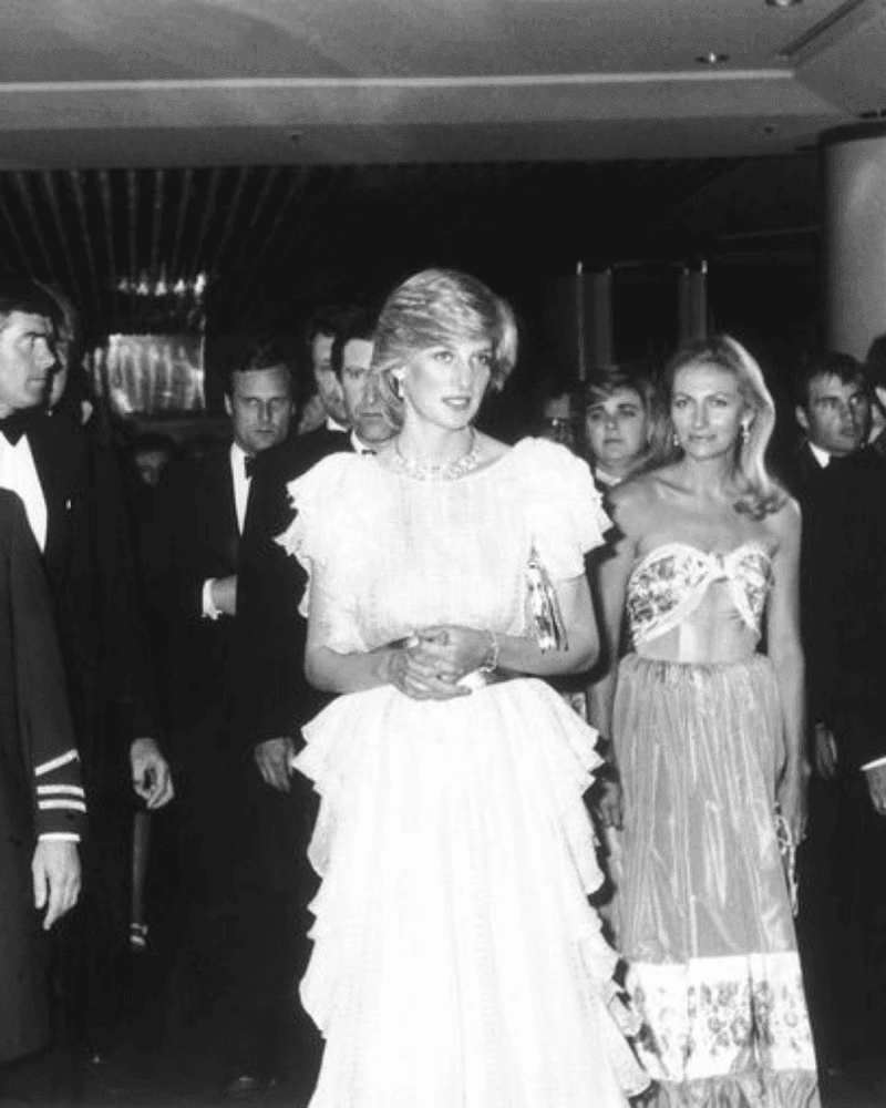 A black and white photo of Princess Diana and another woman walking among people in formal attire.