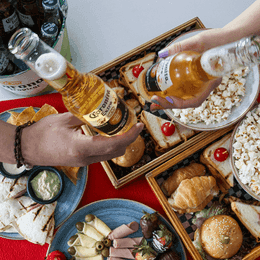 Hands clinking beer bottles above a breakfast spread with pastries, popcorn, and fruit at Unika Hotels, Restaurant Bogota