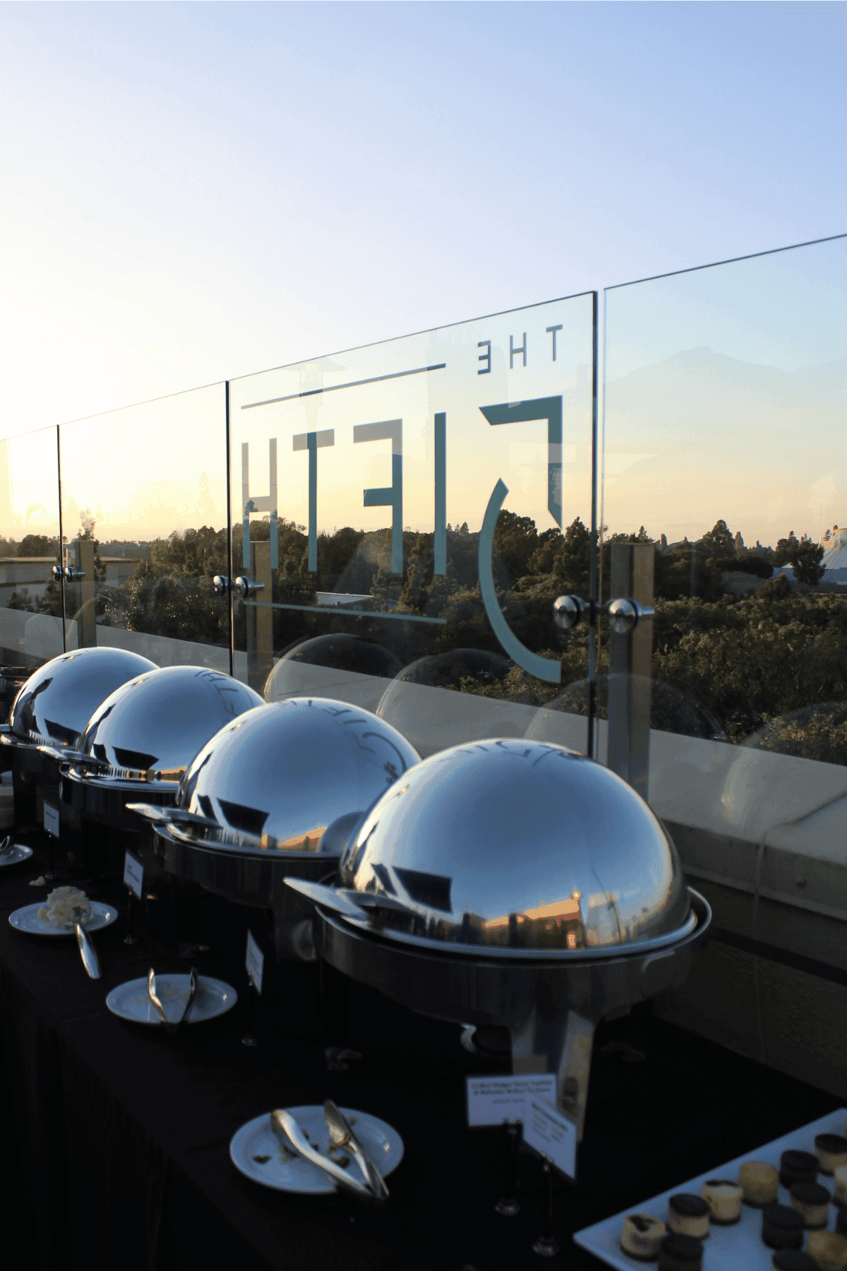 A buffet-style dining area with silver chafing dishes on a black tablecloth, overlooking a scenic view at sunset.
