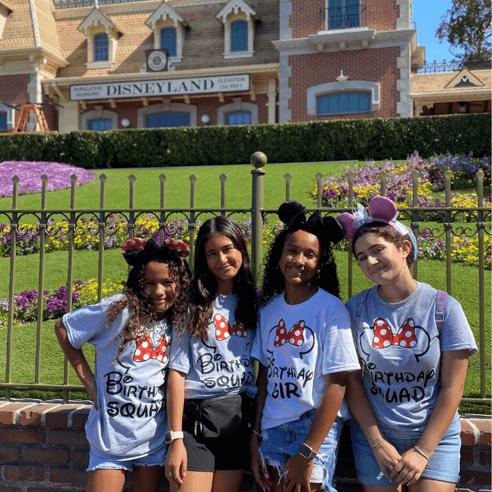 Four friends in matching birthday squad shirts pose in front of the iconic Disneyland clock tower entrance on a sunny day.