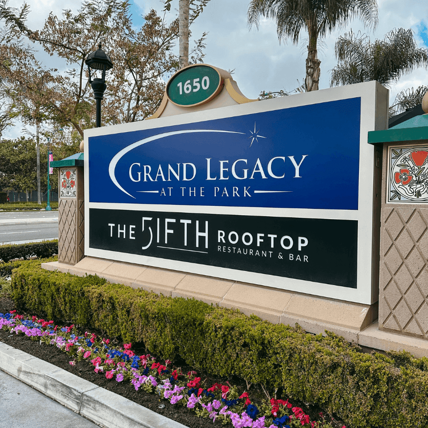 Exterior sign for Grand Legacy at the Park hotel with address 1650 and The 5ifth Rooftop Restaurant & Bar, surrounded by flowers.