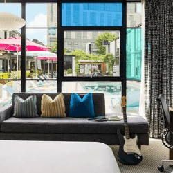Modern hotel room with dark sofa, decorative pillows, and large windows overlooking a pool area with pink umbrellas.