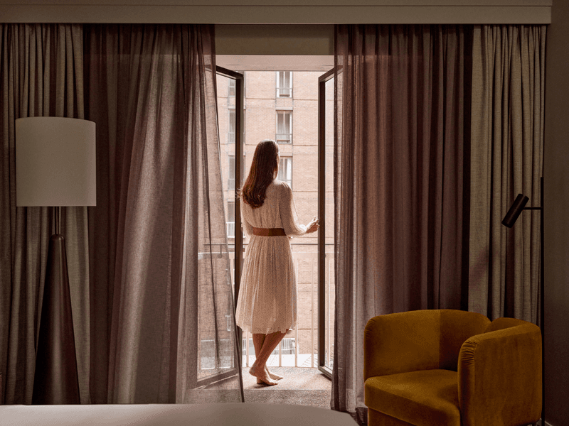Woman in a dress stands on a balcony overlooking a building from a hotel room.