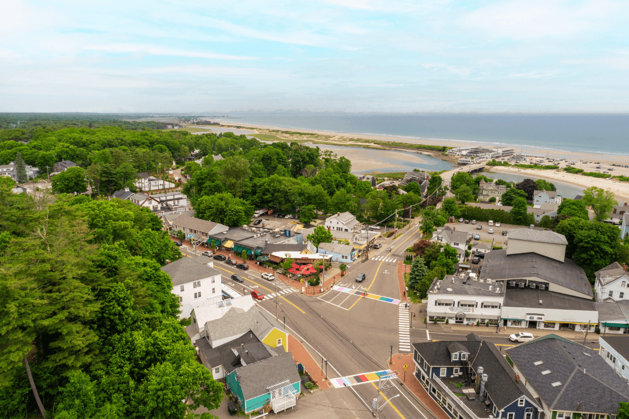 An aerial view of a coastal town with buildings, trees, and a beach in the background.