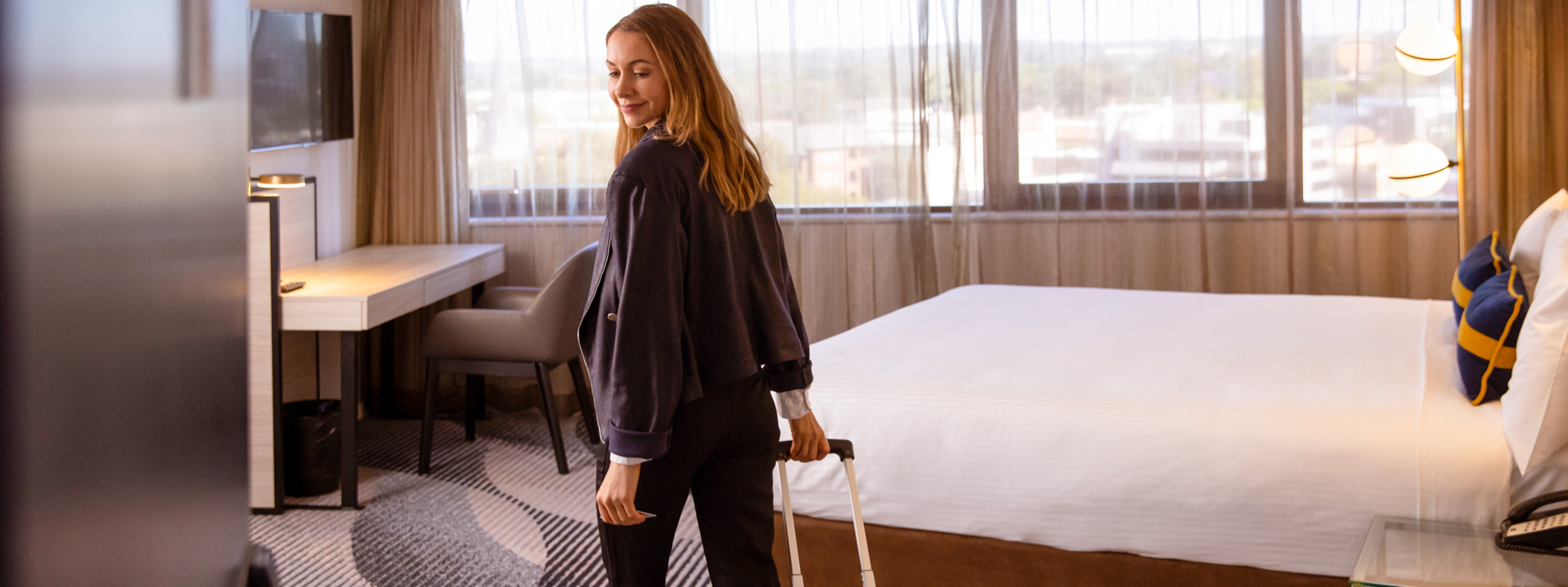 Guest by a suitcase in a Standard Queen Room at Novotel Sydney Parramatta, with a bed and a work desk under orb lights