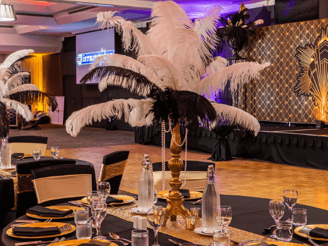 Banquet hall with tables, chairs, and large feather centerpieces, illuminated by purple and gold lights.