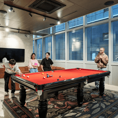 Four people playing pool on a red table in a large room with windows and a carpet.