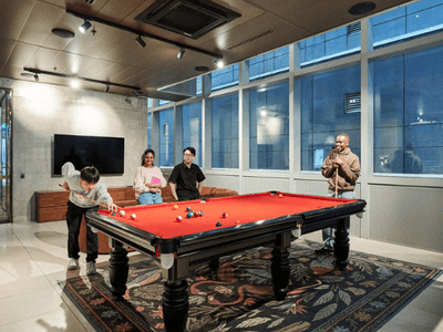 Four people playing pool on a red table in a large room with windows and a carpet.