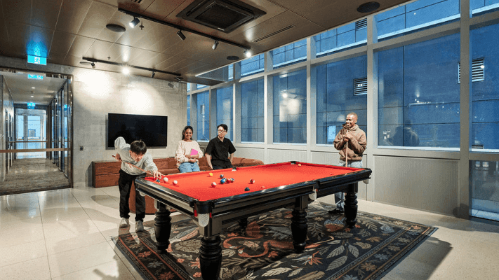 Four people playing pool on a red table in a large room with windows and a carpet.