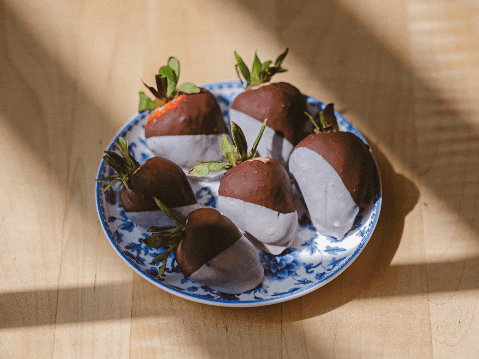 Chocolate-covered strawberries with white stripes on a blue and white patterned plate on a wooden table.