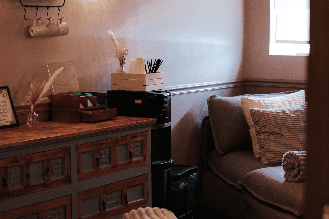 Coffee station with mugs and paper cups by a wooden dresser beside a cushioned lounge chair at La Tourelle Inn and Spa