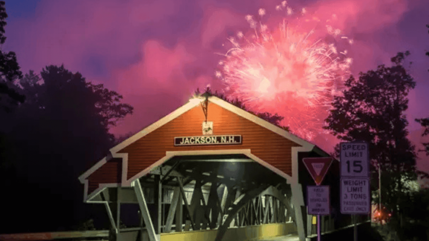 Jackson, N.H. covered bridge with fireworks display and speed limit 15 sign at night.