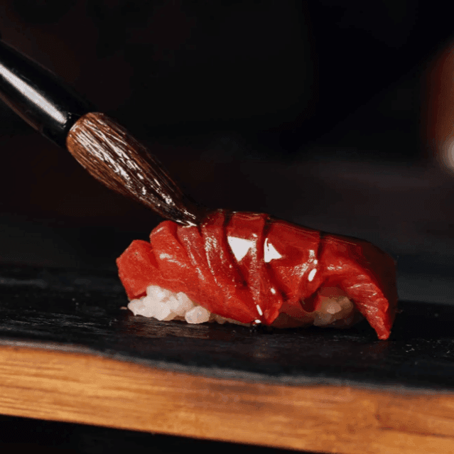 A brush glazes fresh tuna sushi on a black slate plate during an omakase experience.