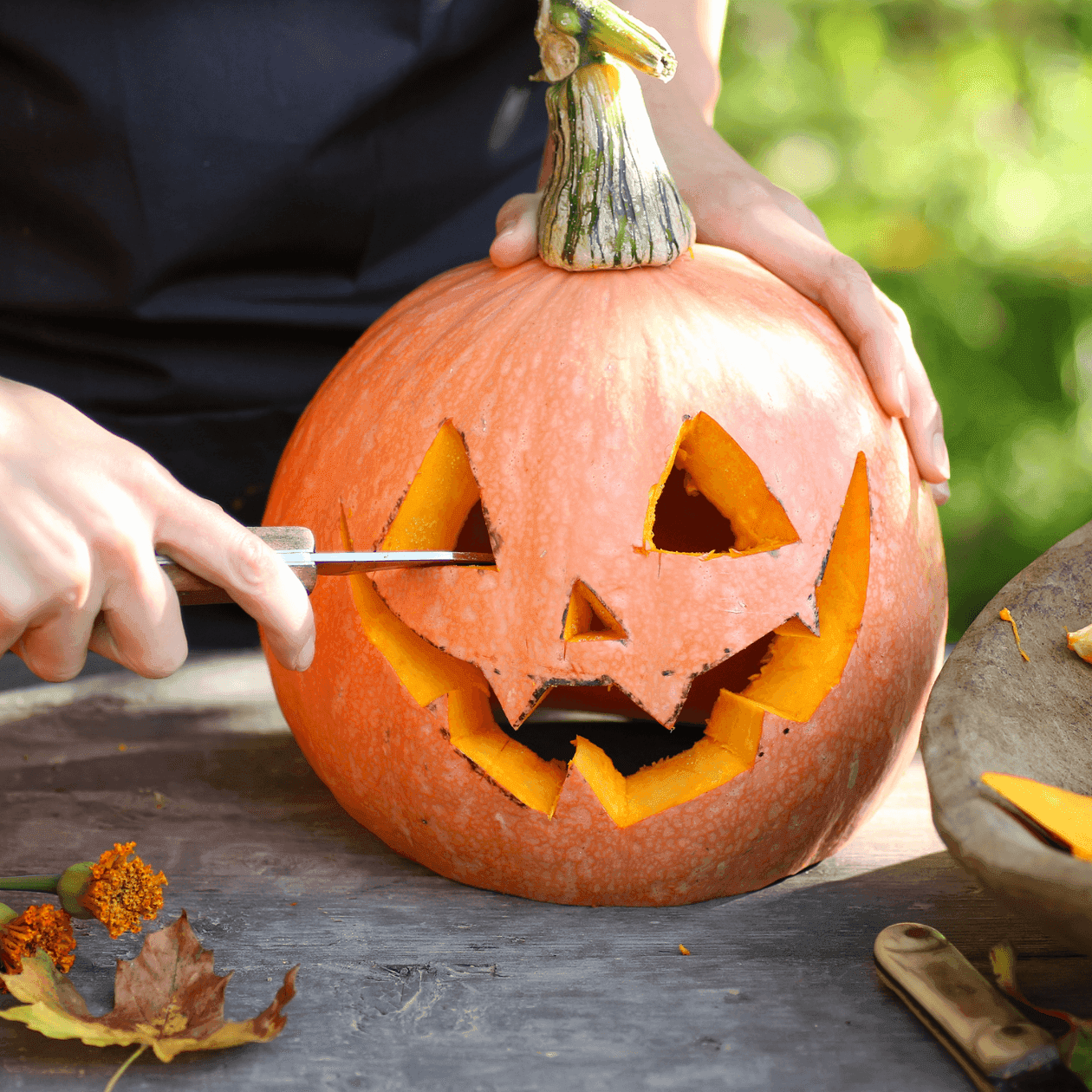 Person carving a pumpkin with a knife, bowl of pumpkin seeds, and flowers on a table.