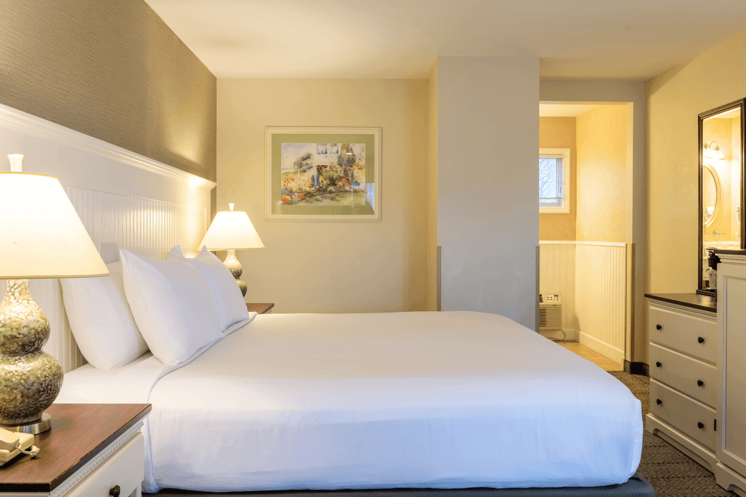 A hotel room with a large bed, two lamps, a painting on the wall, and a doorway to the bathroom.